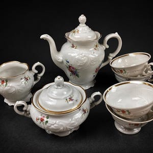 May include: A white porcelain tea set with floral accents. The set includes a teapot, sugar bowl, creamer, and two teacups with saucers. Gold trim edges the pieces. The set is arranged on a black surface.