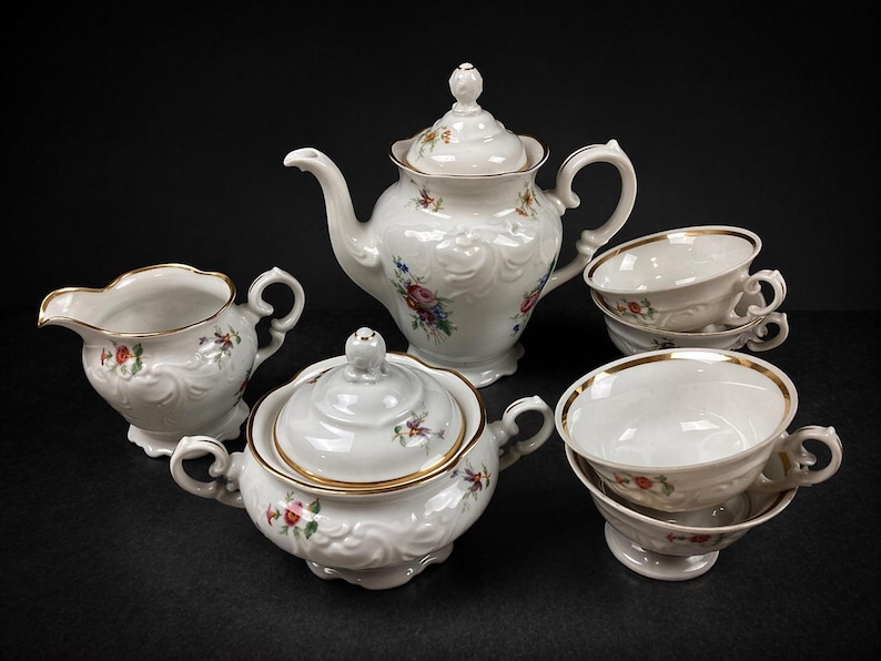 May include: A white porcelain tea set with floral accents. The set includes a teapot, sugar bowl, creamer, and two teacups with saucers. Gold trim edges the pieces. The set is arranged on a black surface.