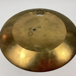 Puede incluir: Un plato grande y redondo de lat&oacute;n con un acabado ligeramente deslustrado. La parte posterior del plato presenta un colgador de metal y una peque&ntilde;a pegatina. El plato tiene un dise&ntilde;o circular y un color dorado c&aacute;lido.