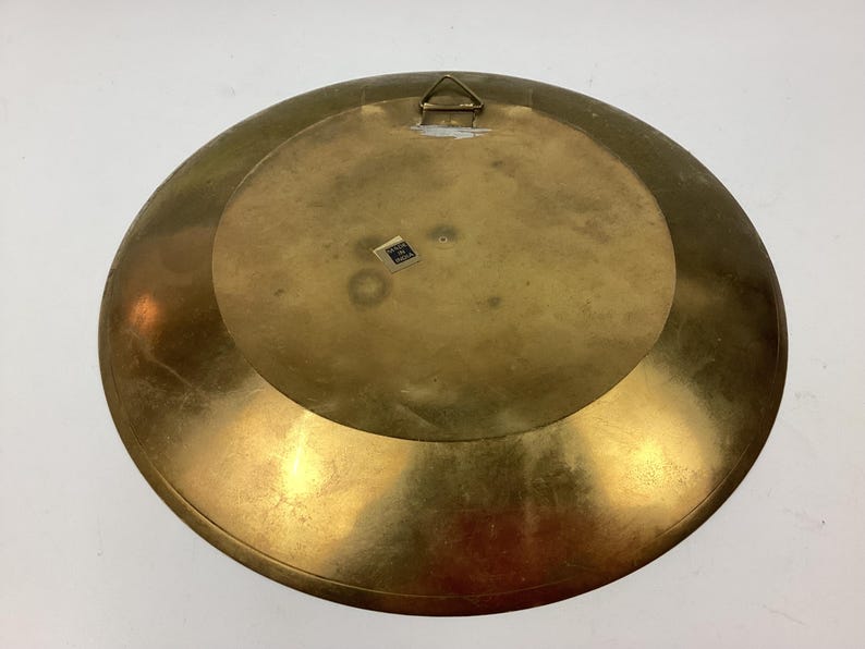 Puede incluir: Un plato grande y redondo de lat&oacute;n con un acabado ligeramente deslustrado. La parte posterior del plato presenta un colgador de metal y una peque&ntilde;a pegatina. El plato tiene un dise&ntilde;o circular y un color dorado c&aacute;lido.