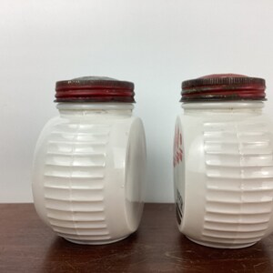 以下が含まれることがあります： 赤色の金属製の蓋が付いた、2つのヴィンテージの白いガラス瓶。瓶の側面にはリブのデザインがあり、空のようです。1つの瓶には側面に赤い文字があります。瓶は茶色の表面に置かれています。