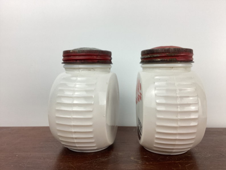 以下が含まれることがあります： 赤色の金属製の蓋が付いた、2つのヴィンテージの白いガラス瓶。瓶の側面にはリブのデザインがあり、空のようです。1つの瓶には側面に赤い文字があります。瓶は茶色の表面に置かれています。