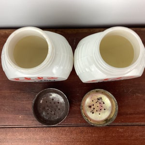 以下が含まれることがあります： 赤色の文字と金属製の蓋が付いた、2つの白いヴィンテージソルトシェーカー。シェーカーはリブのデザインで、開いており、内部が見えます。蓋は外れており、塩を出すための穴が見えています。