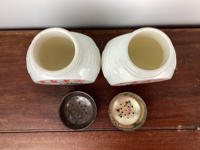 以下が含まれることがあります： 赤色の文字と金属製の蓋が付いた、2つの白いヴィンテージソルトシェーカー。シェーカーはリブのデザインで、開いており、内部が見えます。蓋は外れており、塩を出すための穴が見えています。
