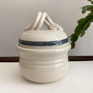 May include: A cream-colored ceramic jar with a matching lid featuring a twisted handle and small decorative circular accents. The lid is adorned with a horizontal dark blue band, and the jar body has subtle horizontal ridges along its exterior.