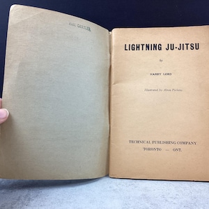 May include: Open vintage book titled "Lightning Ju-Jitsu" by Harry Lord. The book's cover is a light tan color with black text. The book is open, revealing the title page and the inside cover. The book is from the Technical Publishing Company in Toronto, Ontario.