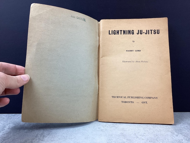May include: Open vintage book titled "Lightning Ju-Jitsu" by Harry Lord. The book's cover is a light tan color with black text. The book is open, revealing the title page and the inside cover. The book is from the Technical Publishing Company in Toronto, Ontario.