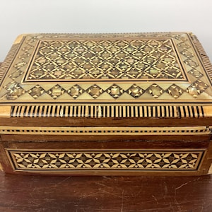 May include: A wooden box with intricate inlaid designs. The top of the box features a geometric pattern of light and dark wood, while the bottom has a more floral design. The box is closed.