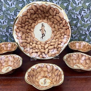 May include: Set of six peanut-shaped bowls with a gold rim. The bowls are decorated with a pattern of peanuts and a cartoon peanut character. The bowls are arranged in a circle around a larger peanut-shaped bowl with the same design. The text "Mr. Peanut" is printed on the larger bowl.