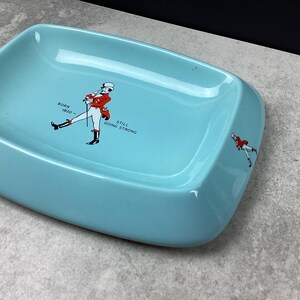 May include: A pale blue rectangular ashtray with a graphic of a man in a red coat and the text "BORN 1820 - STILL GOING STRONG". The ashtray has rounded corners and a smaller version of the graphic on the side.