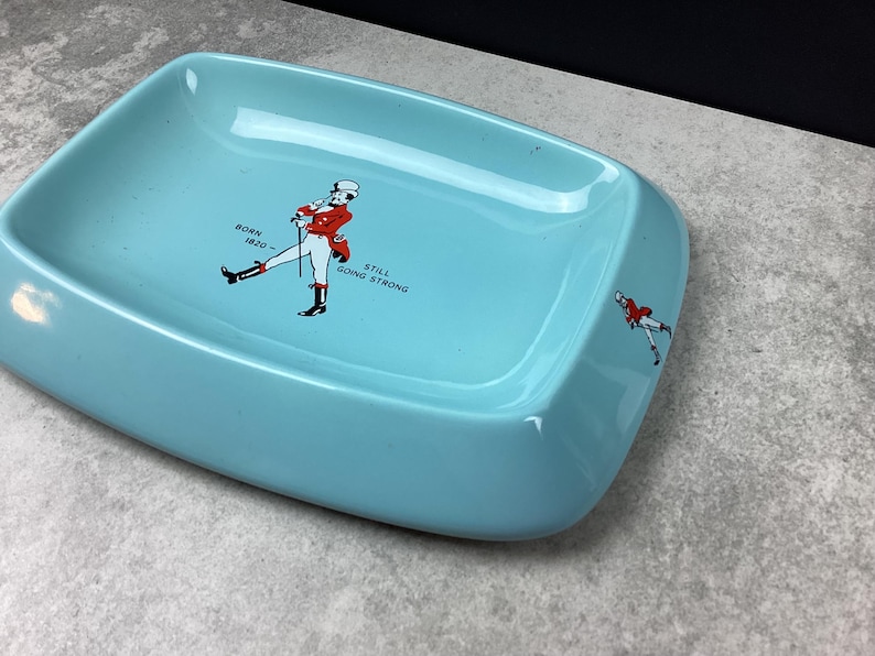 May include: A pale blue rectangular ashtray with a graphic of a man in a red coat and the text "BORN 1820 - STILL GOING STRONG". The ashtray has rounded corners and a smaller version of the graphic on the side.