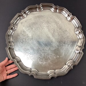 May include: A silver-coloured, ornate serving tray with a scalloped edge. The tray features an engraved floral design and text that reads "V.I. LOMORE ONTARIO HOTEL, NIAGARA". The tray is round and appears to be made of metal.