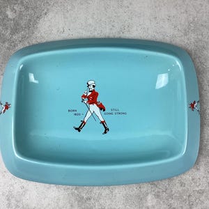 May include: A rectangular, light blue tray with rounded corners. The tray features a graphic of a man in a red coat and black boots, with the text "BORN 1820 - STILL GOING STRONG". Decorative elements are on the sides.