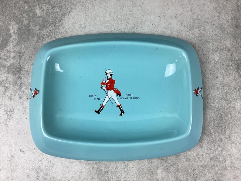 May include: A rectangular, light blue tray with rounded corners. The tray features a graphic of a man in a red coat and black boots, with the text "BORN 1820 - STILL GOING STRONG". Decorative elements are on the sides.