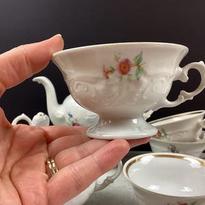 May include: White porcelain teacup with a floral design and ornate handle. The teacup is held in a hand, with other teacups and a teapot visible in the background. The teacup has a pedestal base and a gold rim.