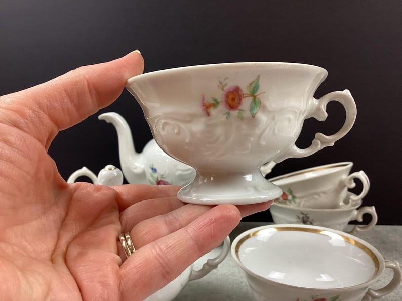May include: White porcelain teacup with a floral design and ornate handle. The teacup is held in a hand, with other teacups and a teapot visible in the background. The teacup has a pedestal base and a gold rim.