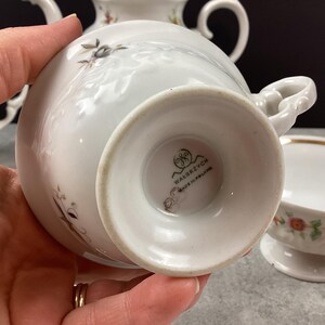 May include: White porcelain teacup with a floral design and gold trim. The teacup is part of a tea set, with a matching saucer and teapot visible in the background. The bottom of the teacup has the text "WAŁBRZYCH MADE IN POLAND".