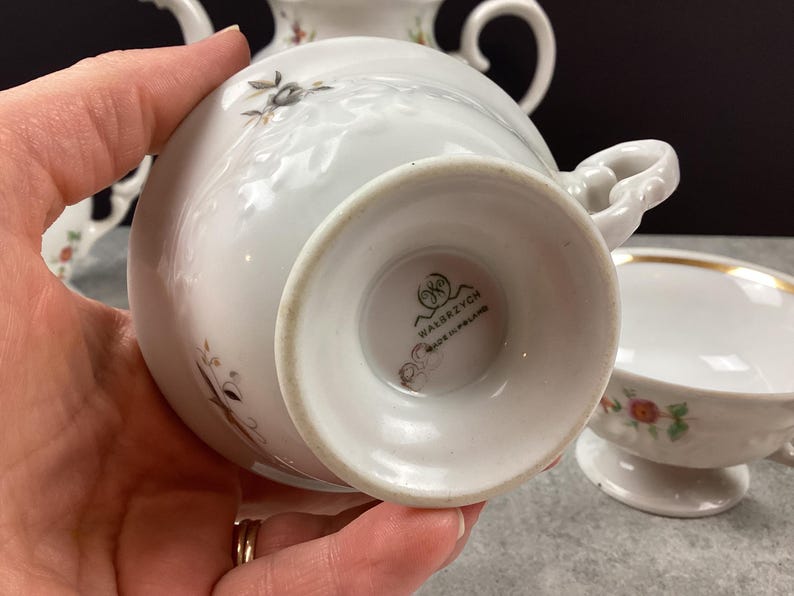 May include: White porcelain teacup with a floral design and gold trim. The teacup is part of a tea set, with a matching saucer and teapot visible in the background. The bottom of the teacup has the text "WAŁBRZYCH MADE IN POLAND".