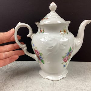 May include: White porcelain teapot with a lid, handle, and spout. The teapot is decorated with floral patterns in pink, blue, and yellow. The rim and the finial on the lid have gold accents. The teapot is on a light gray surface.