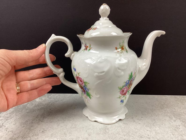 May include: White porcelain teapot with a lid, handle, and spout. The teapot is decorated with floral patterns in pink, blue, and yellow. The rim and the finial on the lid have gold accents. The teapot is on a light gray surface.