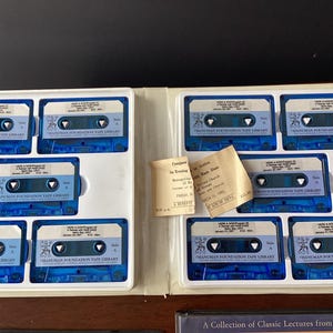 May include: A collection of audio cassette tapes in a blue and white case. The tapes are labeled "Hanuman Foundation Tape Library" and feature a small image of a lion. The case also includes a ticket for a lecture titled "Companions in Loving" by "John Ram Dass".