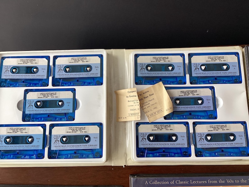 May include: A collection of audio cassette tapes in a blue and white case. The tapes are labeled "Hanuman Foundation Tape Library" and feature a small image of a lion. The case also includes a ticket for a lecture titled "Companions in Loving" by "John Ram Dass".