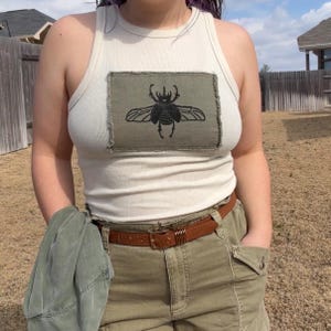 May include: Cream ribbed vest top with an olive green rectangular patch displaying a black beetle design. The person is wearing olive green shorts and a brown belt. A green jacket is held in the left hand.