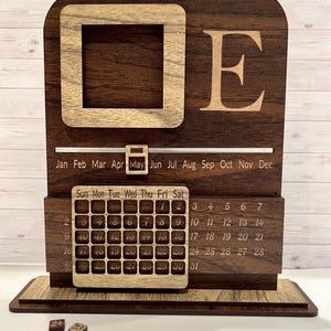 May include: A dark brown wooden perpetual calendar with a light wood square frame and the letter E engraved on the top. It features a sliding month marker currently set to May, and a movable grid overlay for the days of the month, with four small wooden number tiles resting in front.