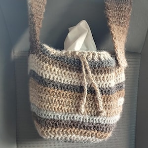 Hanging from a car headrest is a crocheted bag in shades of brown with a drawstring. Inside the bag is a small square tissue box.