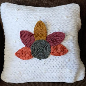 A pillow with a white cover and many white buttons sits on a brown chair. Attached to the pillow is a stylized crocheted image of a turkey (no head) with a brown body and feathers in red, gold, and orange.