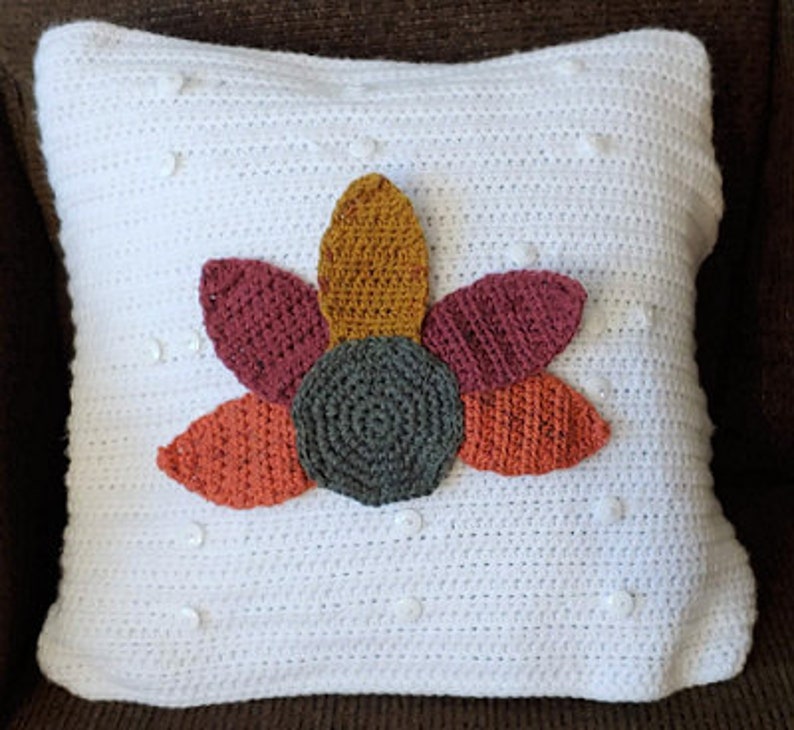 A pillow with a white cover and many white buttons sits on a brown chair. Attached to the pillow is a stylized crocheted image of a turkey (no head) with a brown body and feathers in red, gold, and orange.
