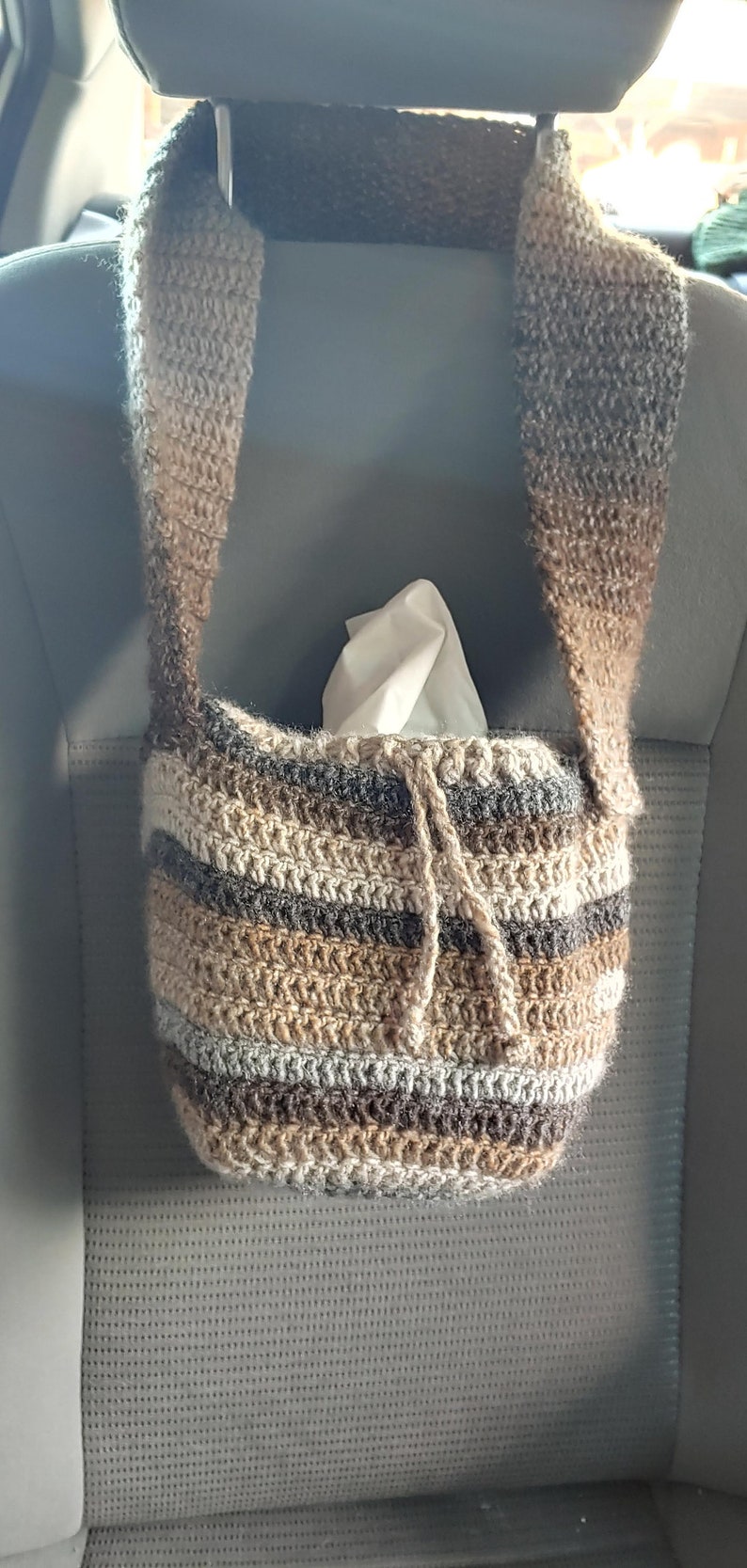 Hanging from a car headrest is a crocheted bag in shades of brown with a drawstring. Inside the bag is a small square tissue box.