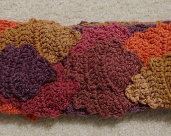 May include: A handmade crochet headband in a fall color palette. The headband features a textured design with leaf-like shapes in shades of orange, brown, burgundy, and purple. The accessory is designed to be worn around the head.