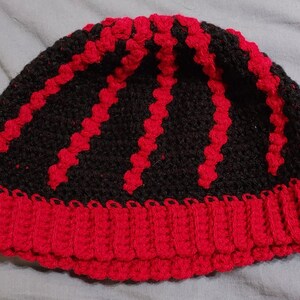 May include: A handmade crochet beanie in black and red. The hat features a ribbed red brim and vertical red stripes against a black background. The hat is a winter accessory.