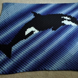 May include: A crocheted blanket featuring a black and white orca whale design on a blue and white gradient background.