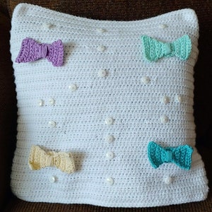 Può includere: Un cuscino quadrato bianco all'uncinetto con quattro fiocchi colorati e bottoni bianchi sparsi. I fiocchi sono viola, verde chiaro, giallo e turchese. Il cuscino è posto su una superficie marrone.
