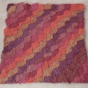 Puede incluir: Una manta de ganchillo hecha a mano en tonos cálidos de rosa, naranja y marrón. La manta presenta un patrón texturizado en forma de escamas. Sus dimensiones son de aproximadamente 127 cm por 152 cm, ofreciendo un diseño visualmente atractivo. Ideal para la decoración del hogar.