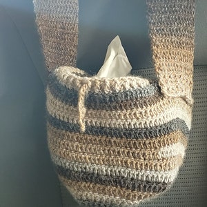 Hanging from a car headrest is a crocheted bag in shades of brown with a drawstring. Inside the bag is a small square tissue box.