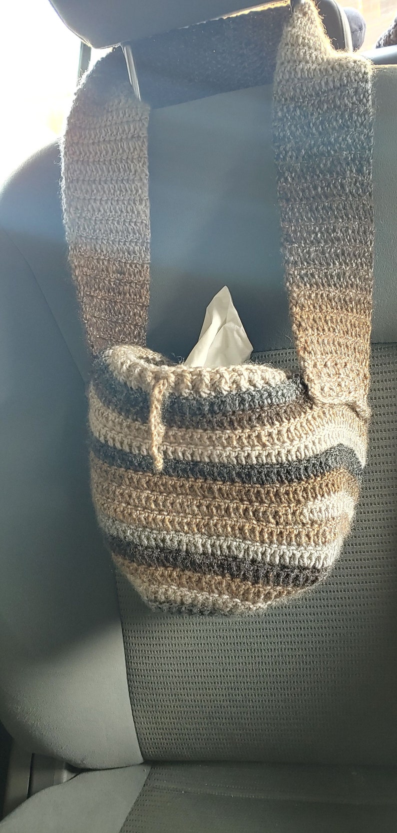 Hanging from a car headrest is a crocheted bag in shades of brown with a drawstring. Inside the bag is a small square tissue box.
