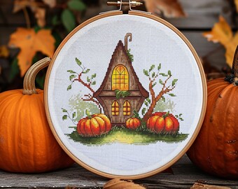 May include: A cross-stitch embroidery in a wooden hoop depicts a whimsical brown house with a glowing yellow window, surrounded by orange pumpkins and green foliage. The embroidery is set against a backdrop of autumn leaves and pumpkins.