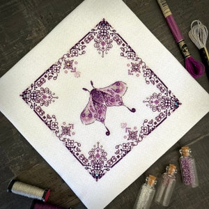 Puede incluir: Un diseño de bordado de punto de cruz morado y blanco con una polilla con detalles intrincados y un borde geométrico.
