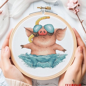 Puede incluir: Patrón de punto de cruz de un cerdo con gafas de buceo y un snorkel, nadando en agua azul. El patrón está en un aro de bordado de madera.