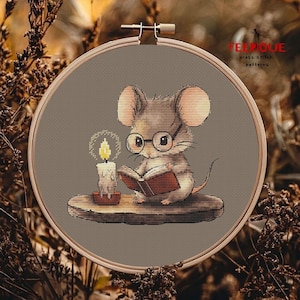 May include: A cross stitch pattern featuring a cute brown mouse wearing glasses and reading a book by candlelight. The mouse is sitting on a wooden ledge. The pattern is in a hoop and ready to be stitched.