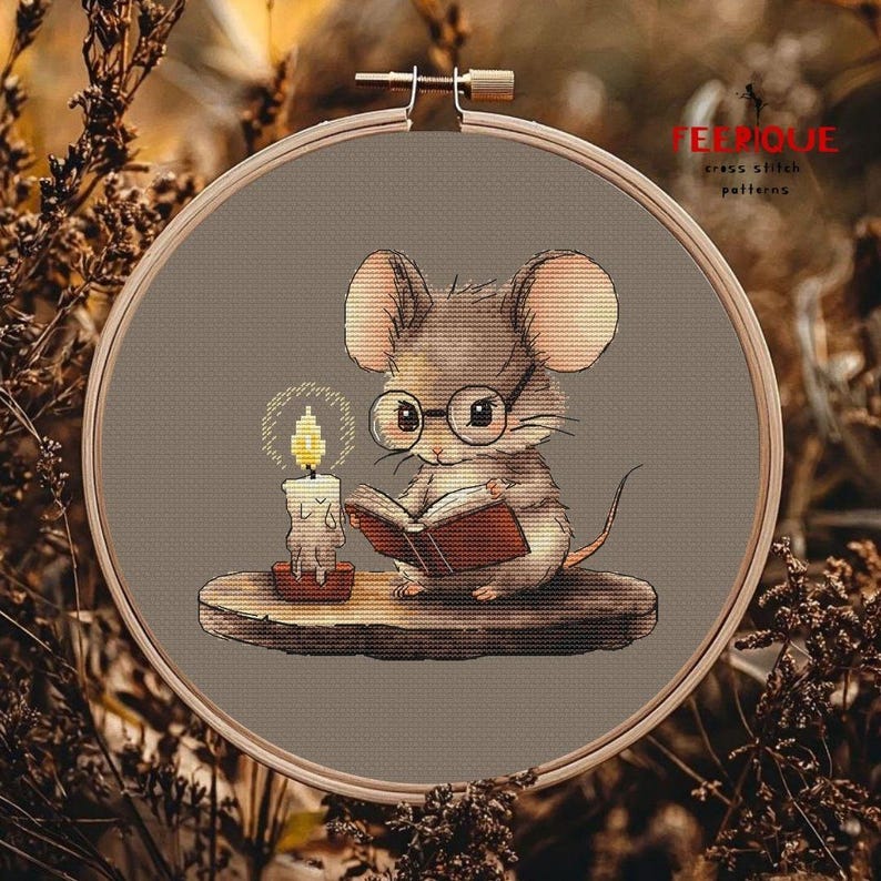 May include: A cross stitch pattern featuring a cute brown mouse wearing glasses and reading a book by candlelight. The mouse is sitting on a wooden ledge. The pattern is in a hoop and ready to be stitched.