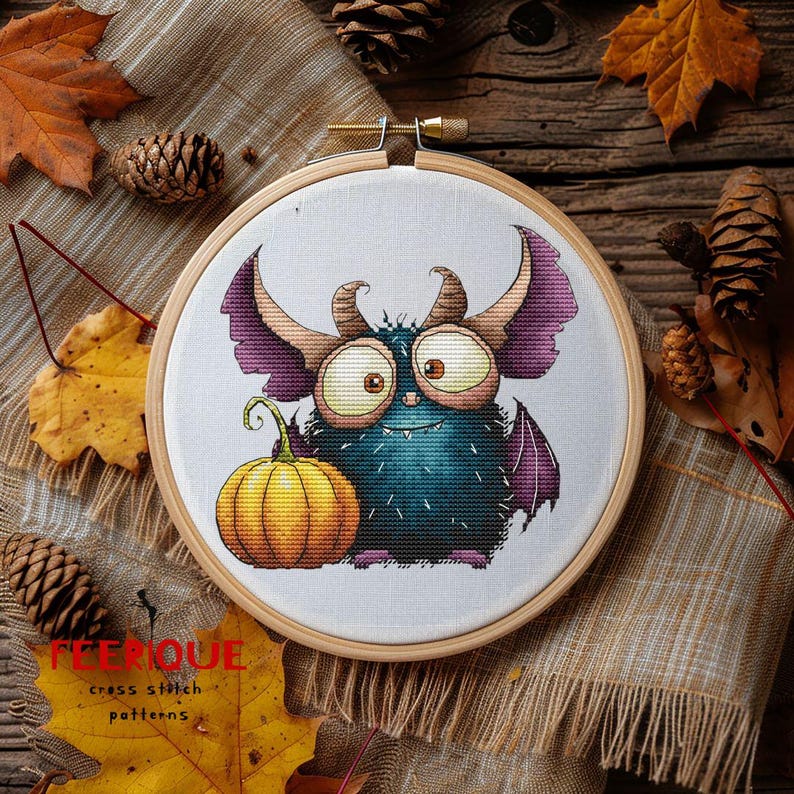 Peut inclure: Un motif de point de croix dans un cercle en bois repr&eacute;sente un monstre bleu sarcelle avec de grands yeux, des cornes et des ailes violettes, &agrave; c&ocirc;t&eacute; d'une citrouille orange. Le motif est sur fond blanc, avec le texte "FEEERIQUE cross stitch patterns" en bas.