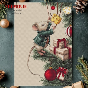 Peut inclure: Illustration fantaisiste d'une souris en veste verte décorant un sapin de Noël avec du fromage et des ornements. L'image comprend le texte "TEERIQUE cross stitch patterns" en rouge, avec des cadeaux et des branches de pin.