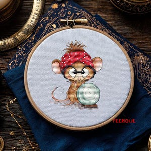 Puede incluir: Bordado de punto de cruz de un ratón de dibujos animados con un turbante de lunares rojos y gafas, mirando una bola de cristal. El bordado está en un aro de madera, con la palabra "FEERIQUE". El fondo incluye una tela azul oscuro y objetos decorativos.