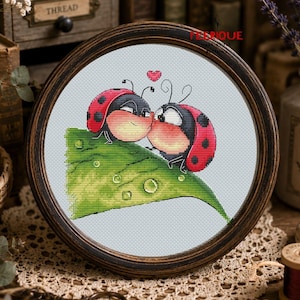 May include: A circular wooden frame containing a cross-stitch embroidery of two red ladybugs with black spots kissing on a green leaf. The leaf features small water droplets, and a small red heart is stitched above the ladybugs. The text FEERIQUE is visible.