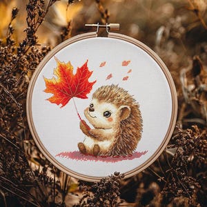 May include: A cute cartoon hedgehog holding a red and orange maple leaf. The hedgehog is sitting on a white background with a few red and orange leaves scattered around it.