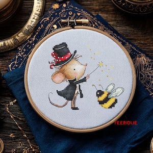 May include: Cross-stitch embroidery in a wooden hoop depicts a mouse magician in a black coat and top hat, using a wand on a yellow and black bee. The design is on light grey fabric, with the word "FEERIQUE" in red.